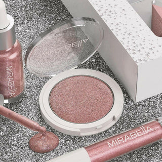 Illuminizing makeup set pieces on a glittery silver background showing the sets highlighting powder up close next to the glow drops & lip gloss.